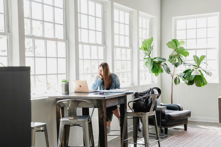 A woman working alone on a laptop in a room with large, bright windows